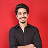 A young man in a red shirt smiling warmly at the camera, photographed against a blurred dark background.