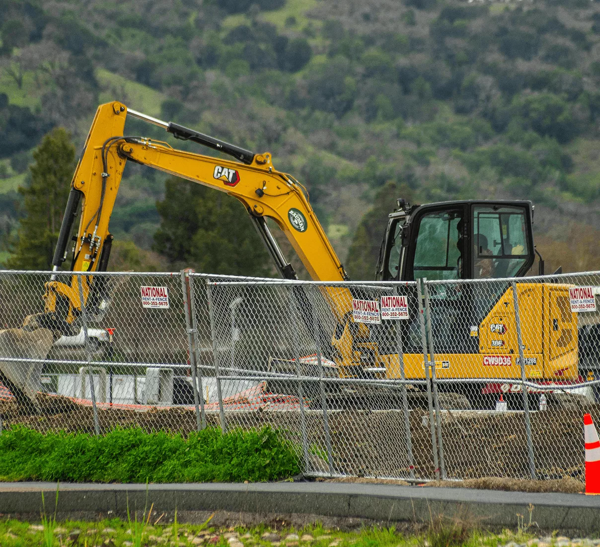 A yellow CAT excavator operating behind a chain-link construction fence with warning signs on a hillside site.