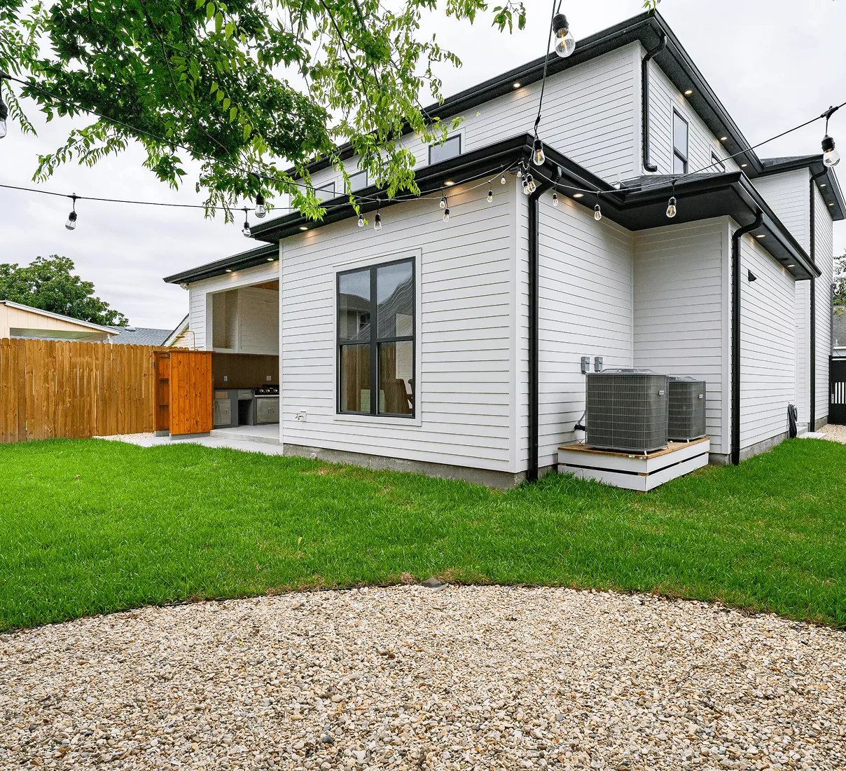 Rear view of a modern white farmhouse with a wooden privacy fence, string lights, and a manicured green lawn.