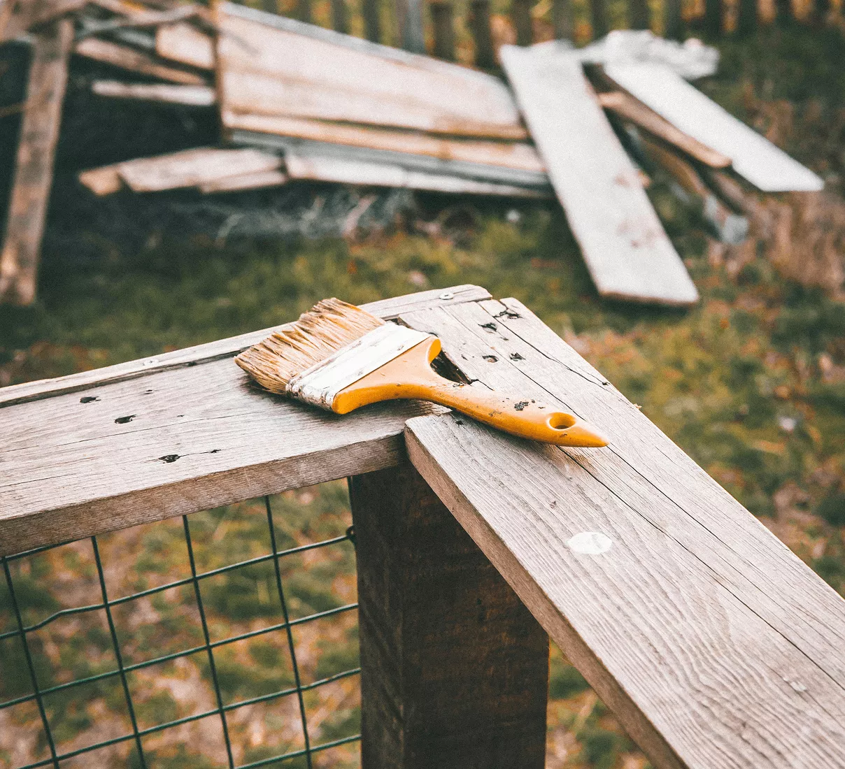 A paint-stained yellow brush resting on a weathered wooden fence rail with scrap lumber piled in the background.