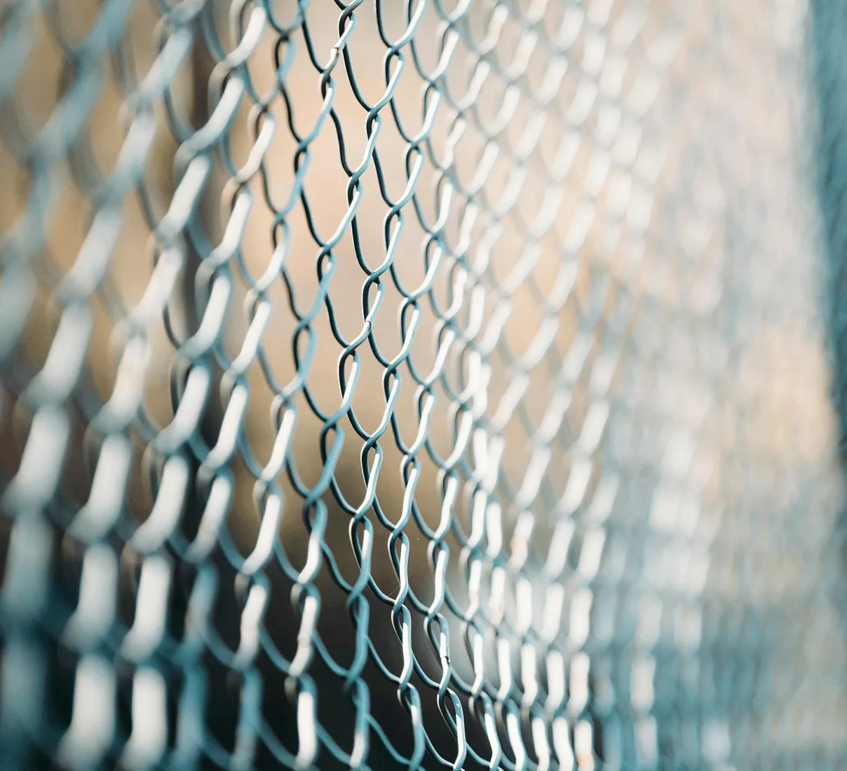 Extreme close-up of a teal-coated steel chain-link fence with a blurred warm-toned background in soft focus.