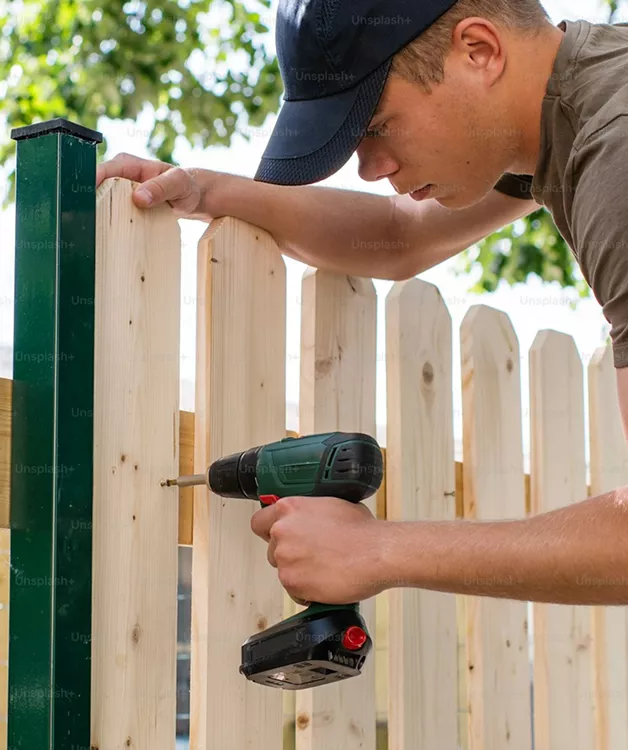 A worker in a cap uses a cordless drill to install wooden picket fence boards onto a green metal post outdoors.