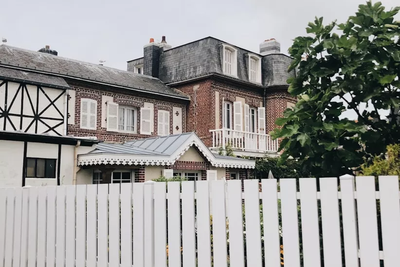 A white picket fence in front of a classic French brick manor house with shuttered windows on an overcast day.