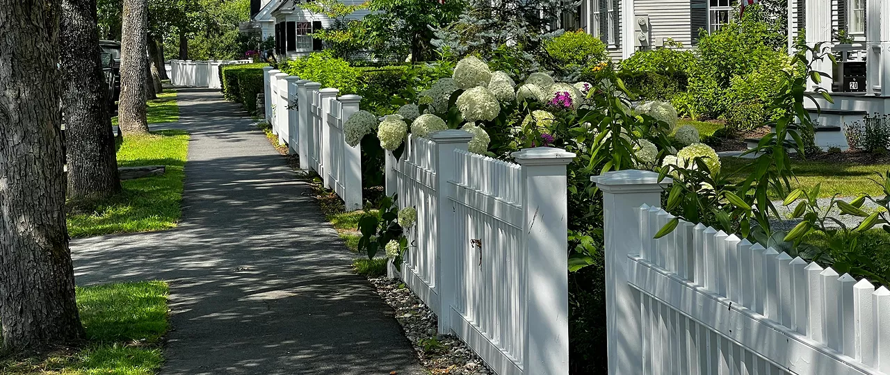 A tree-lined sidewalk beside a white vinyl panel fence with decorative posts and blooming hydrangea bushes in summer.
