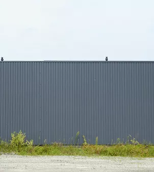 Exterior view of a tall dark gray corrugated steel panel wall against an overcast sky with small trees below.