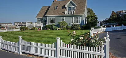 A charming Cape Cod style home with a white picket fence, manicured lawn, and rose bushes on a clear sunny day.