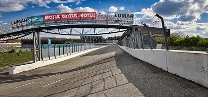 Wide-angle view of a motor racing circuit pit lane with overhead sponsor banners and steel safety barrier fencing.