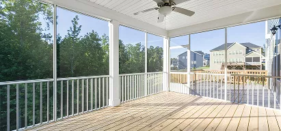 Screened porch with white railing, ceiling fan, and large windows overlooking a tree-lined backyard on a sunny day.