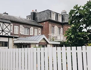 A white picket fence in front of a classic French brick manor house with shuttered windows on an overcast day.