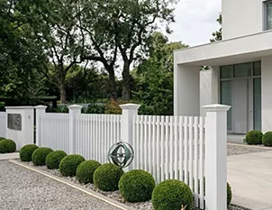 A white picket fence with square posts and trimmed boxwood balls lining the gravel driveway of a modern white villa.
