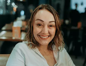 A young woman with wavy hair and a white shirt smiling warmly at the camera in a softly lit indoor setting.
