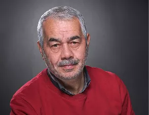 An older man with gray hair and a mustache wearing a red sweater, photographed against a neutral gray background.