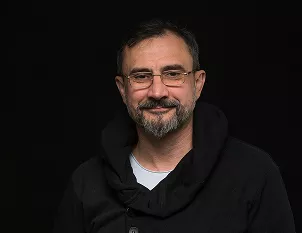 A middle-aged man with a beard and glasses wearing a black hoodie, smiling against a dark studio background.