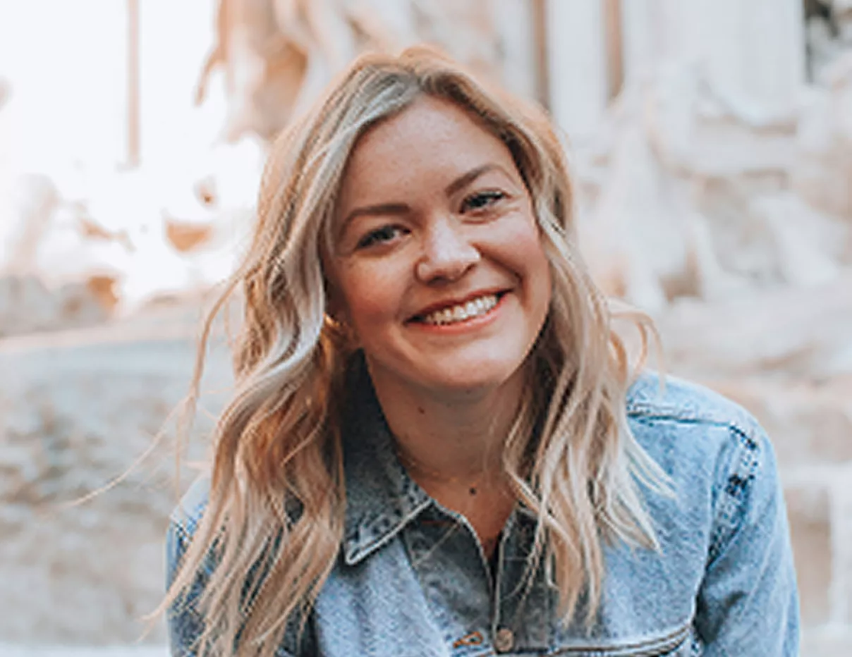 A young woman with long wavy hair wearing a plaid shirt smiling warmly in a bright indoor setting.